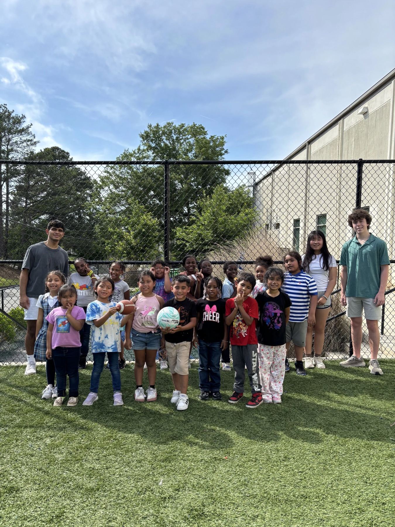 Children holding balls and posing together in front of a chain-link fence outside a community center