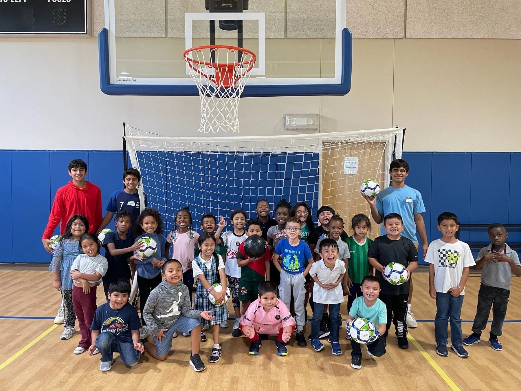 A large group of children sitting and standing together on a gymnasium floor beneath a basketball hoop after a session