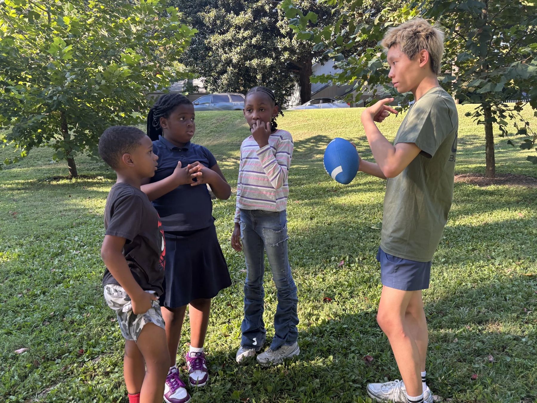 A volunteer holding a ball and talking with children on a sunny grass field