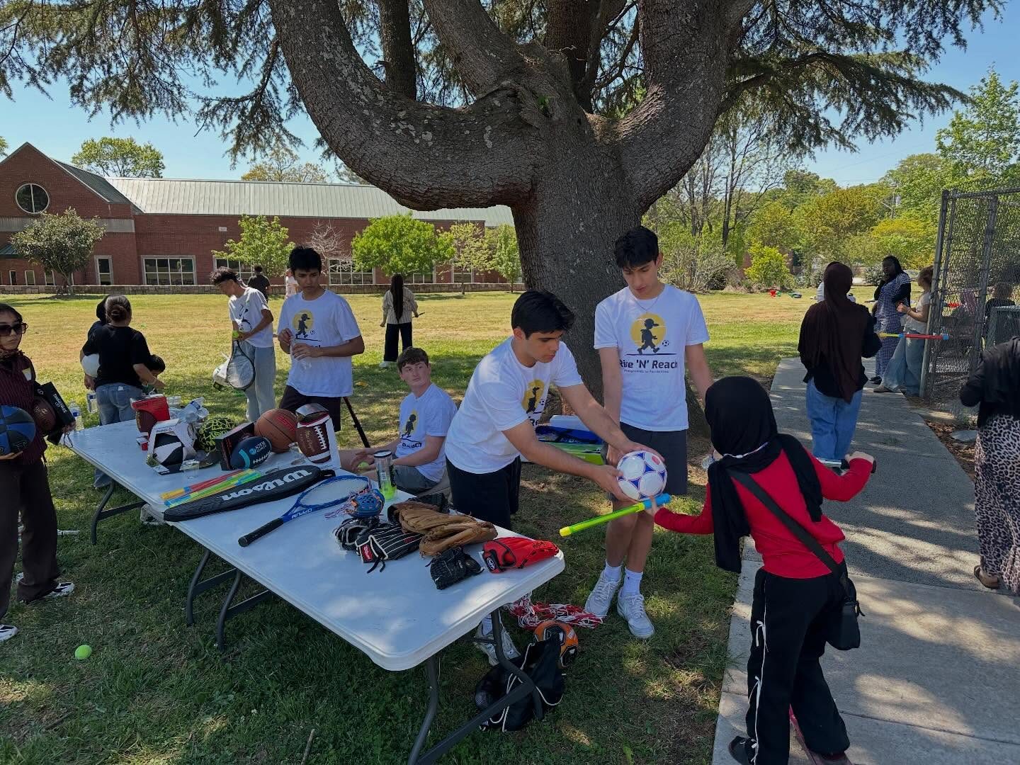 Rise 'N' Reach volunteers in team shirts handing out soccer balls, bats, and gloves to children from a table of donated sports equipment at Clarkston Community Center