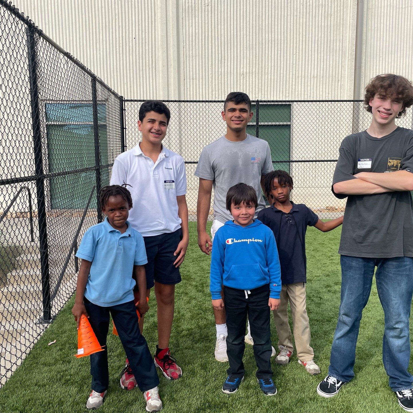 Teen volunteers standing with young children outside beside a chain-link fence on a sunny day